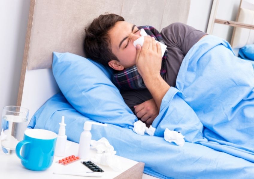 Homem com gripe aparece deitado na cama, enrolado no lençol, com cachecol no pescoço e assoando o nariz num lenço de papel. Na mesinha de cabeceira ao lado da cama, há 1 copo com água, uma xícara e vários medicamentos. 